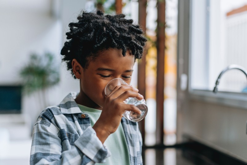 A child with curly dark hair drinks from a glass in a brightly lit kitchen